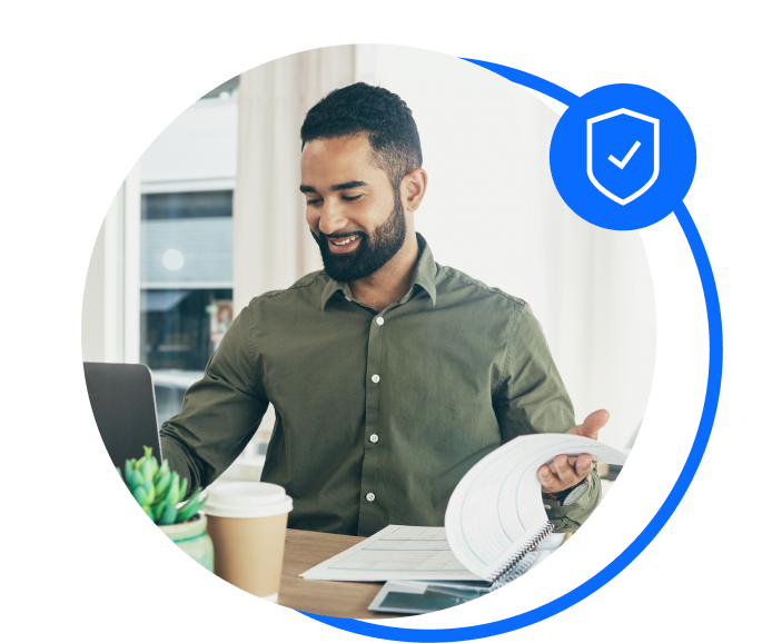 Male bookkeeper wearing a green shirt, sat down at a desk flicking through some paperwork while working on a laptop