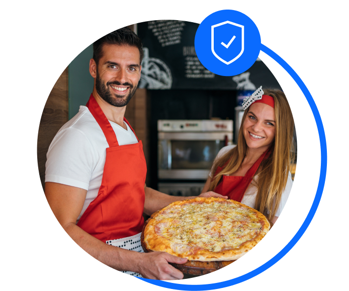 A male and female pizza owner wearing white shirts and red aprons and holding a margarita pizza and smiling at the camera