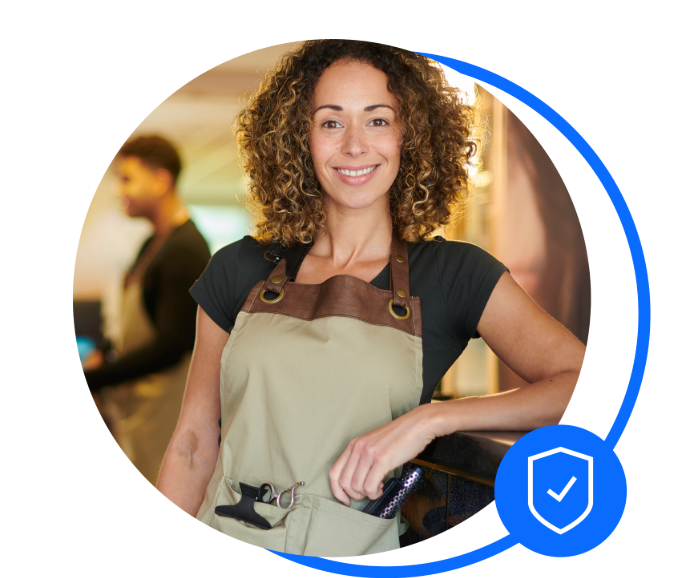 Female mobile hairdresser with brunette girls is standing with one arm leaning on a counter smiling at the camera. She's wearing an apron with hair tools in the pockets - a hair brush, and clips