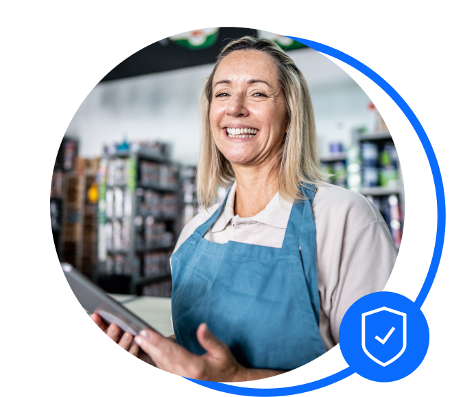Blonde female retailer stood in front of shelves of stock. She's wearing a white t-shirt and blue overalls. She's holding a tablet and smiling at the camera