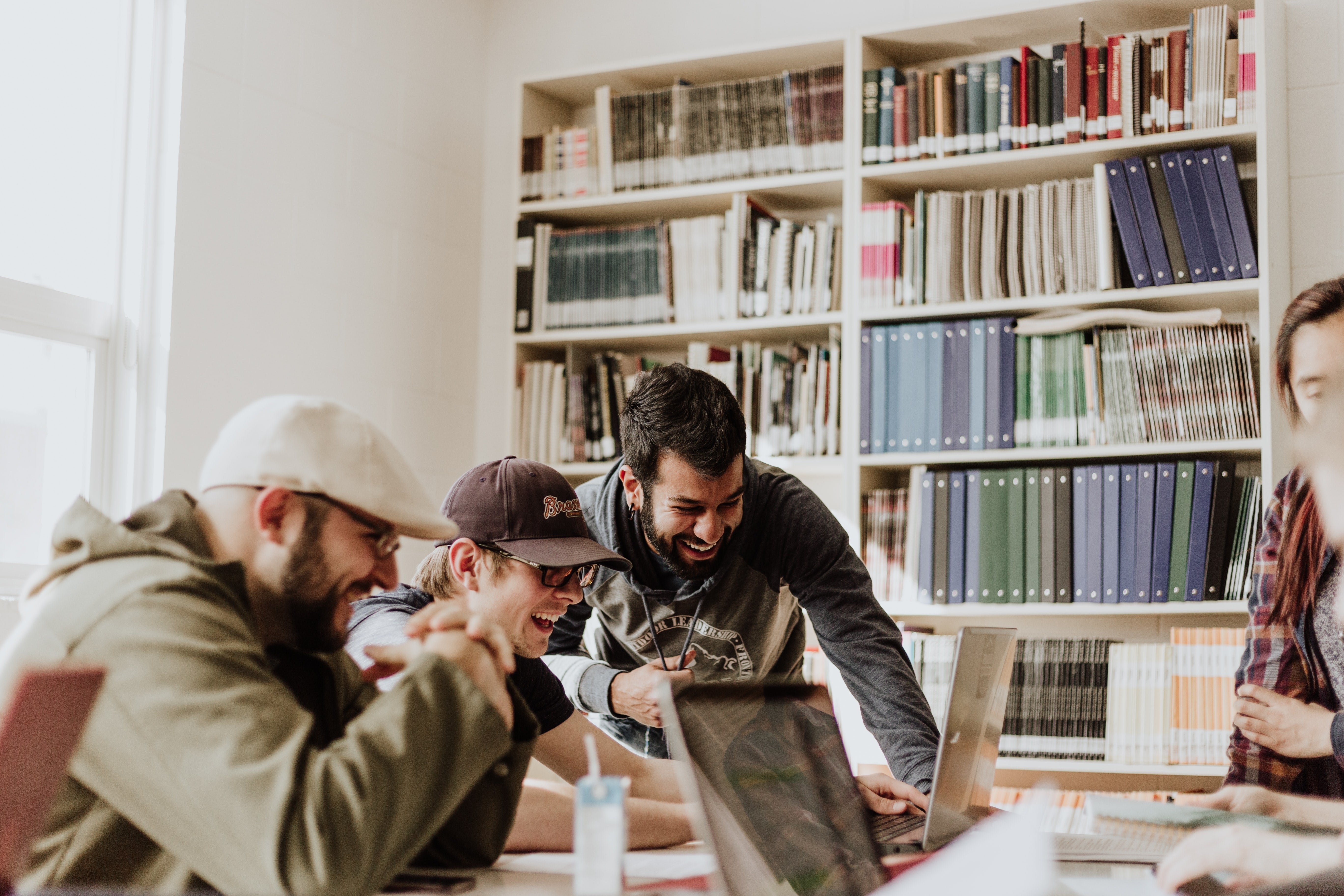 A group of workers at a a tech start up gather around a laptop in their office. Shelves full of books and folders sit in the background.