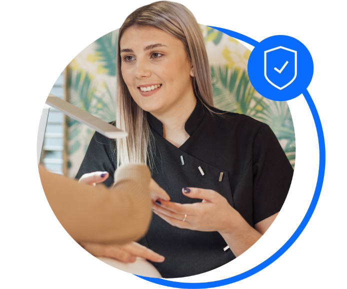 A female beautician sat down and holding a customer's fingers after a nail treatment. She's wearing a black beautician's uniform and has long blonde hair. She's smiling at the camera