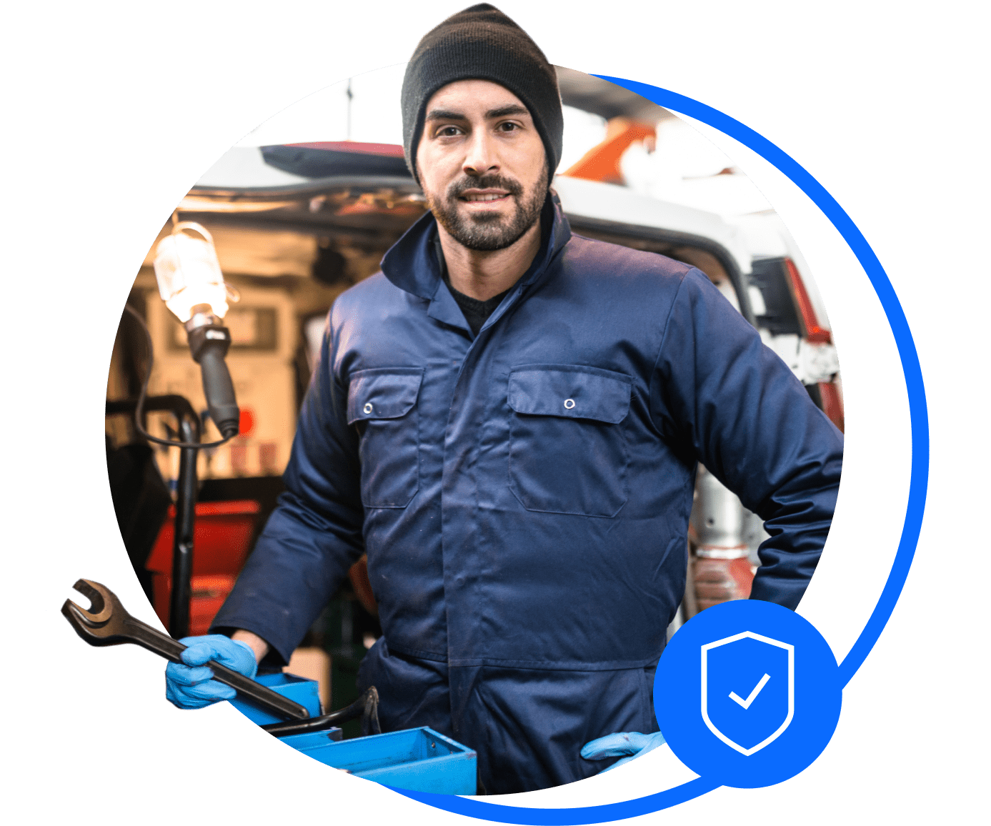 A mechanin in blue overalls and a black hat stands outside his workshop holding a spanner. He's thinking about sorting his workshop insurance.