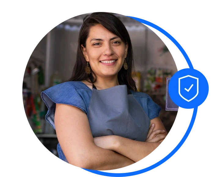 Female with long dark hair and wearing a blue blouse with a blue apron over the top. She's standing facing the camera with her arms folder and smiling