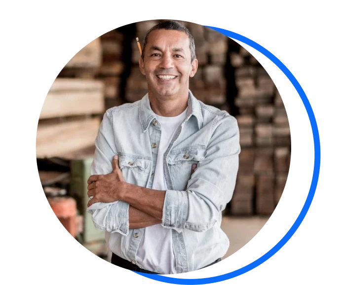 Image of  male carpenter stood cross armed in front of a pile of wood. He has a blue over shirt on and a pencil sitting on his right ear.