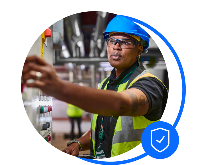 Male engineer with a blue hard hat on and florescent jacket on is stood working next to a wall panel of switches