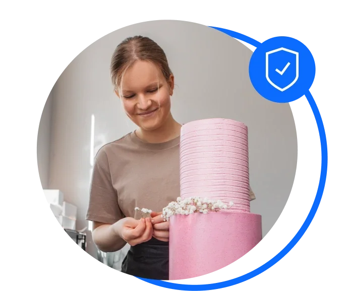 Blonde female in a brown t-shirt stood decorating a two-tier pink cake 
