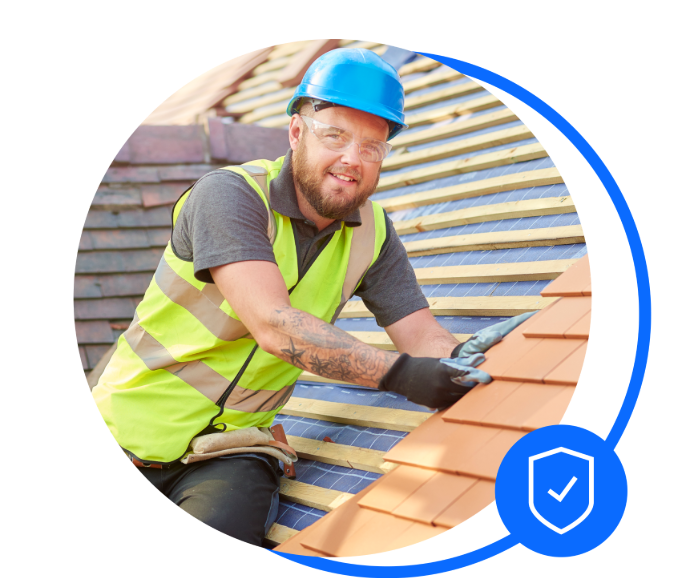 Male in a grey t-shirt, blue hard hat and visibility jacket is sat on a roof placing roof tiles. He's smiling at the camera
