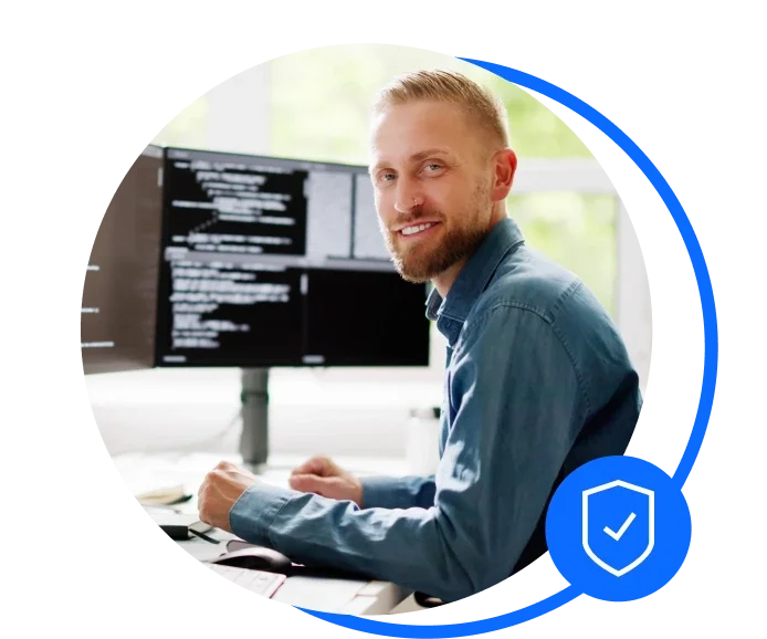 Male IT contractor sat at a desk with two computer screens. He's wearing a blue shirt and smiling at the camera
