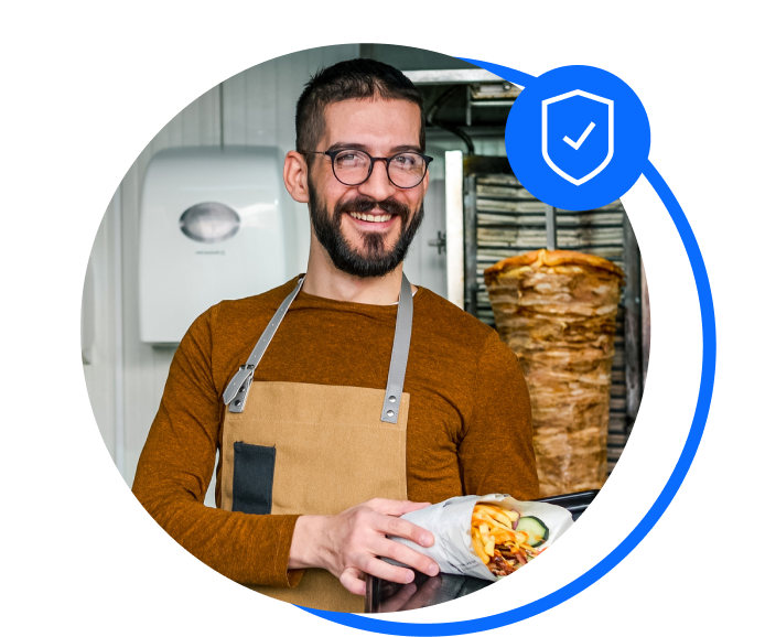 Male kebab shop worker is stood holding a rolled up kebab with chips and salon. He has dark hair and glasses and is wearing a brown long-sleeved t-shirt and a beige apron. He's standing in front of kebab meat being cooked.