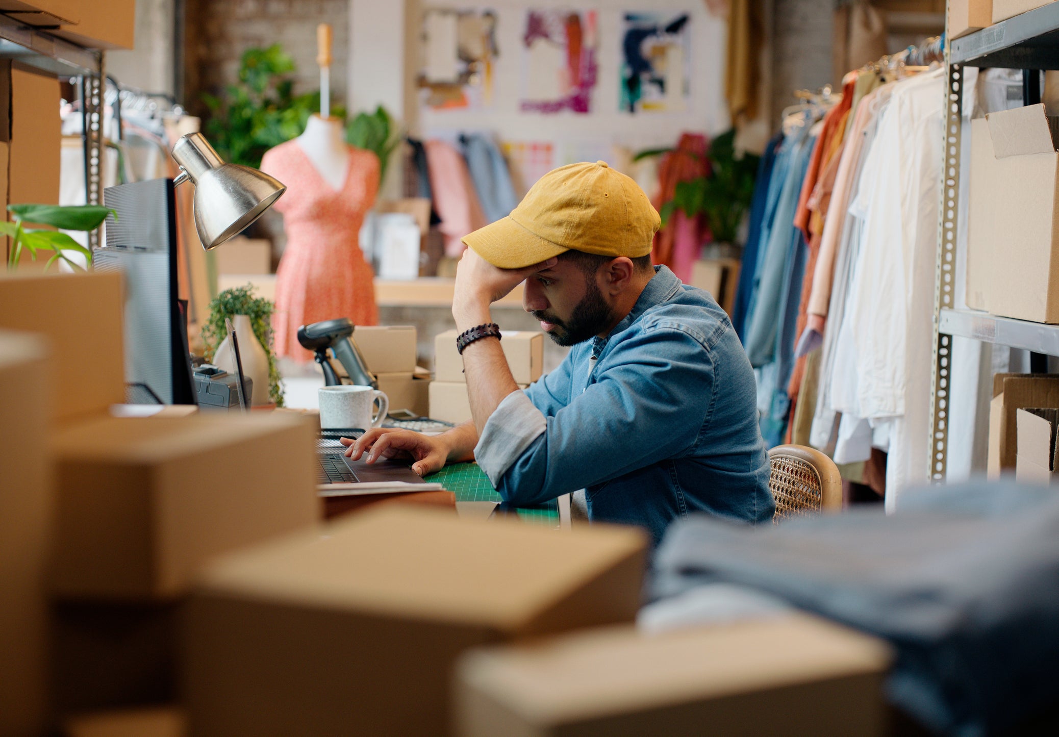 A business owner sits in his stockroom surrounded by clothes and boxes. He's working on his laptop and looks frustrated.