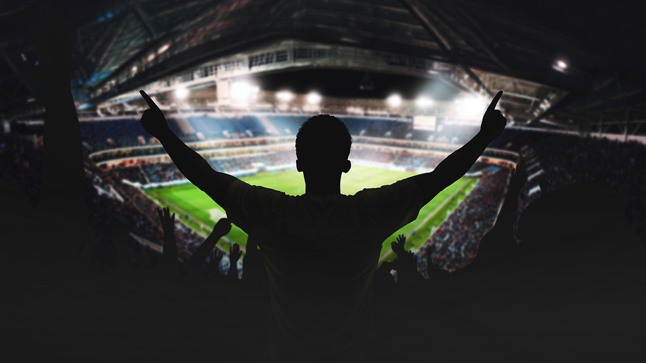 A football fan holding his arms out wide in silhouette in the corner of a football stadium