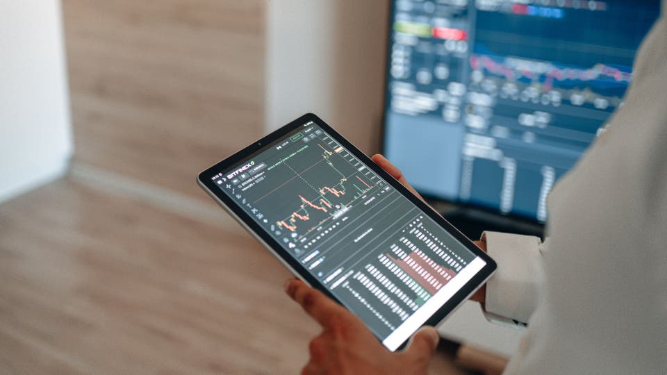 A person holding a tablet with a graph and other technical information on it. In the background is a computer screen with similar information