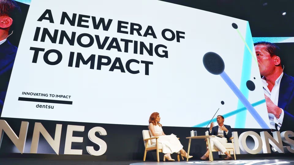 a man and a woman sitting on a stage in conversation with a screen saying 'A New Era Of Innovating To Impact' behind them
