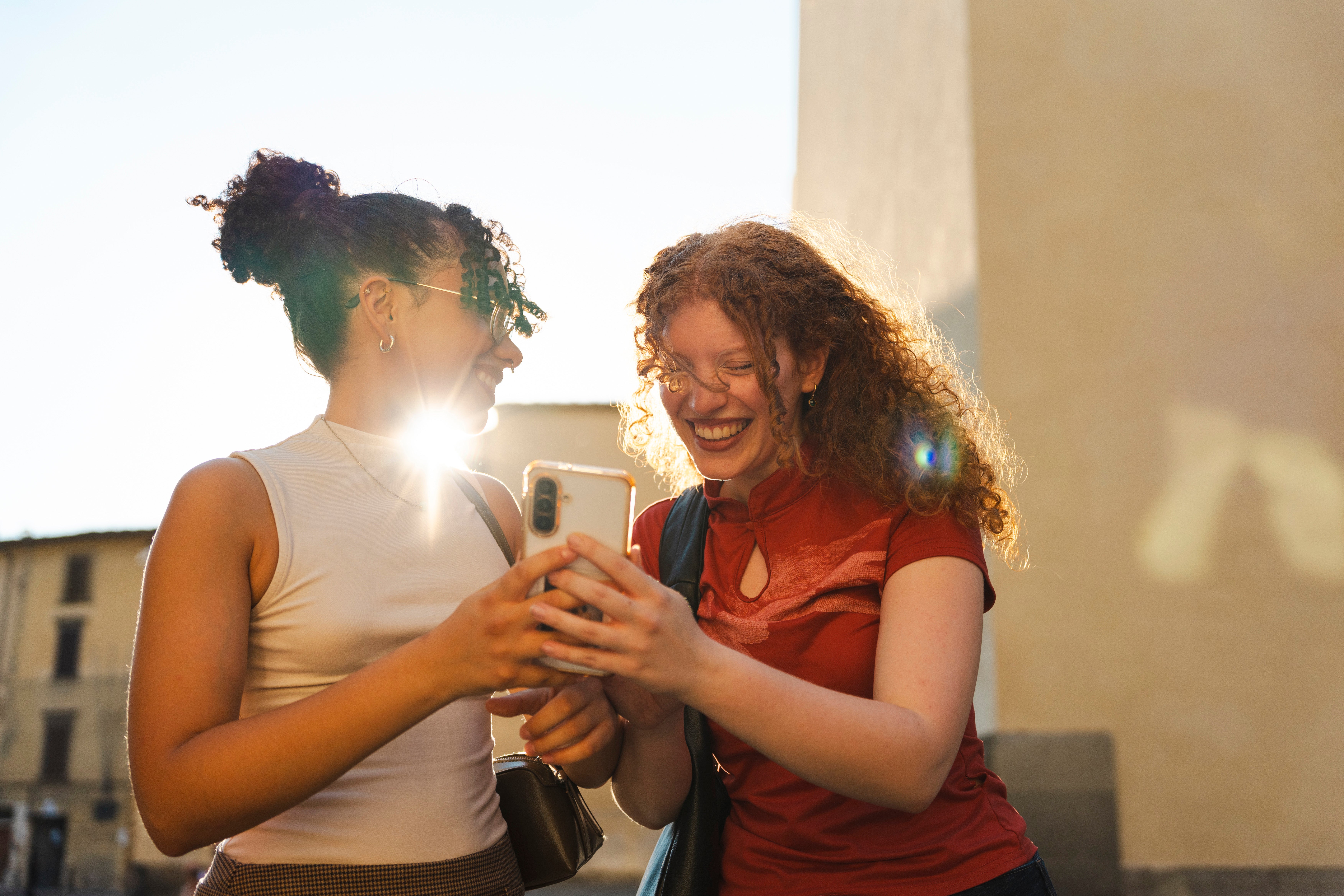 Two women looking at a phone and laugh. The sun is shining between them