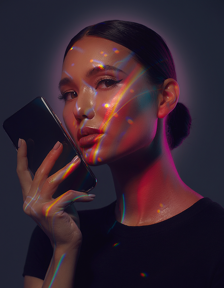 Woman holding a phone by her face. There is a rainbow and other light effects across her face