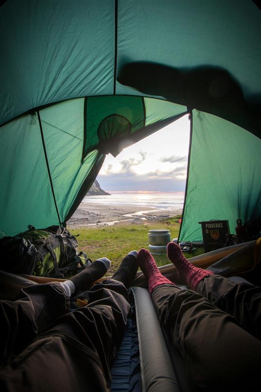 A view from inside a tent looking at a beach and the sea. There are two pairs of legs and a rucksack inside the tent
