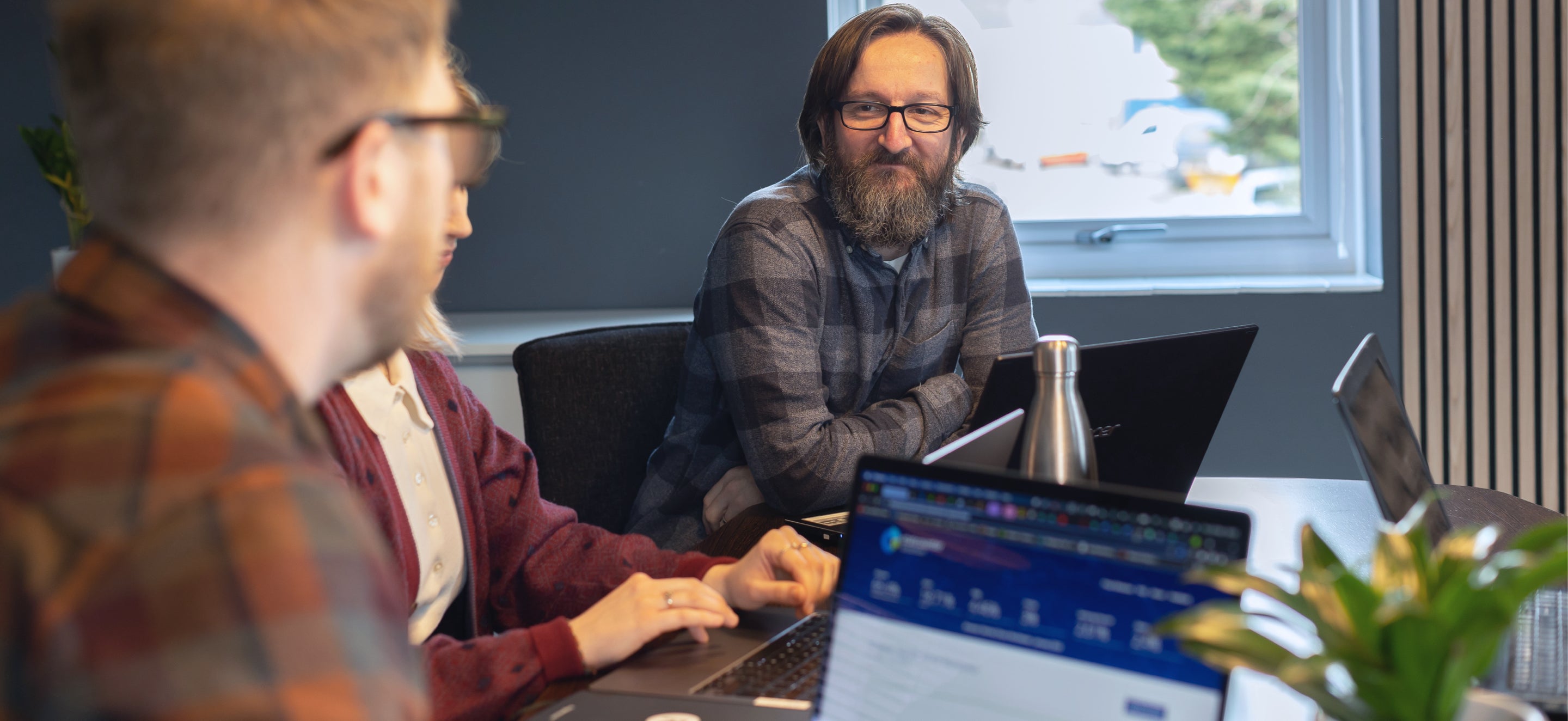 Two men and lady in a meeting room, plus laptops and a window in the background