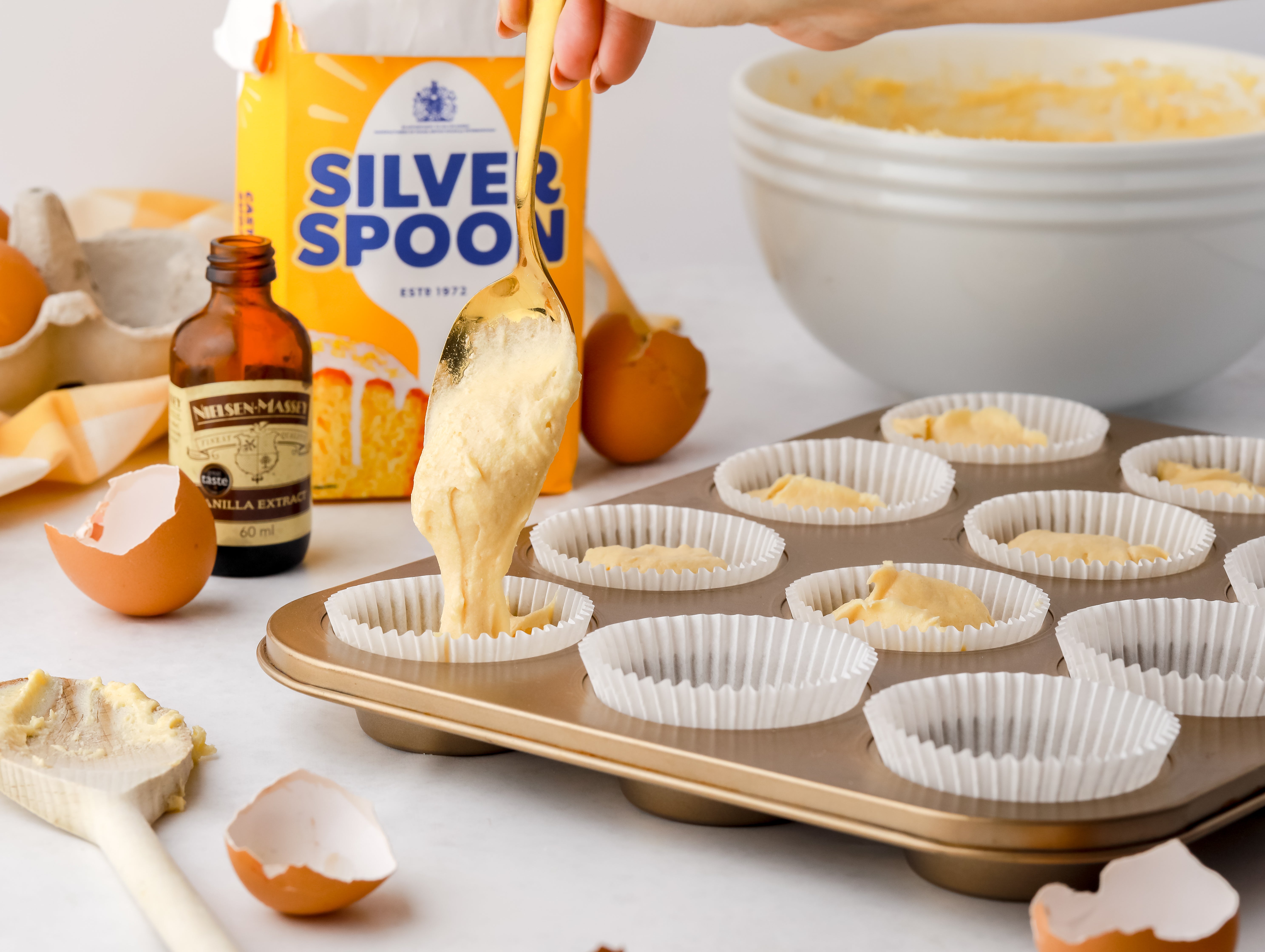 Cake batter being added into cupcake cases. Cracked egg shells, sugar pack and vanilla extract bottle beside.
