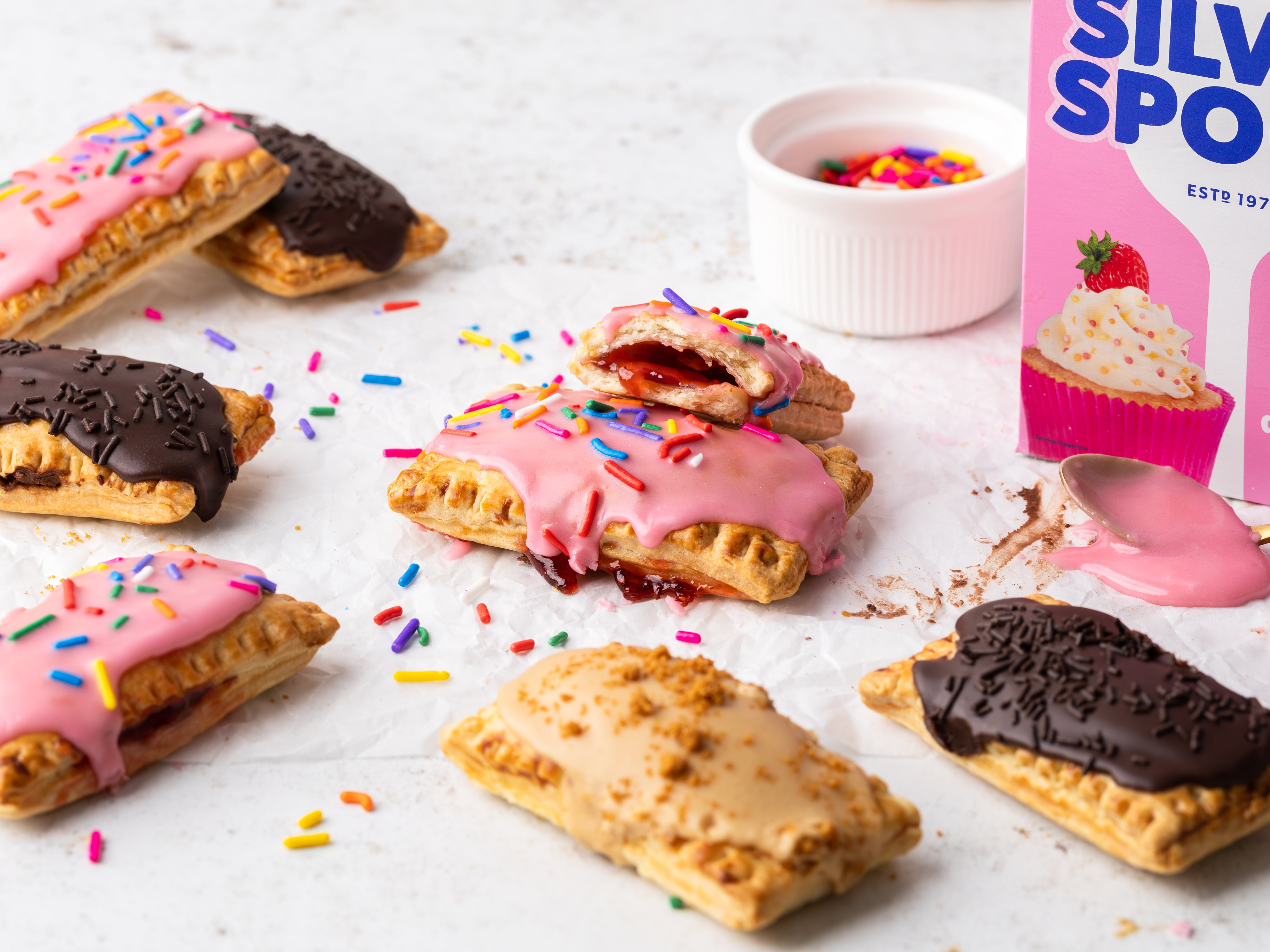 Freshly made Air Fryer Pop Tarts on a marble surface decorated with icing and multi coloured sprinkles next to a pink pack of Silver Spoon icing sugar.
