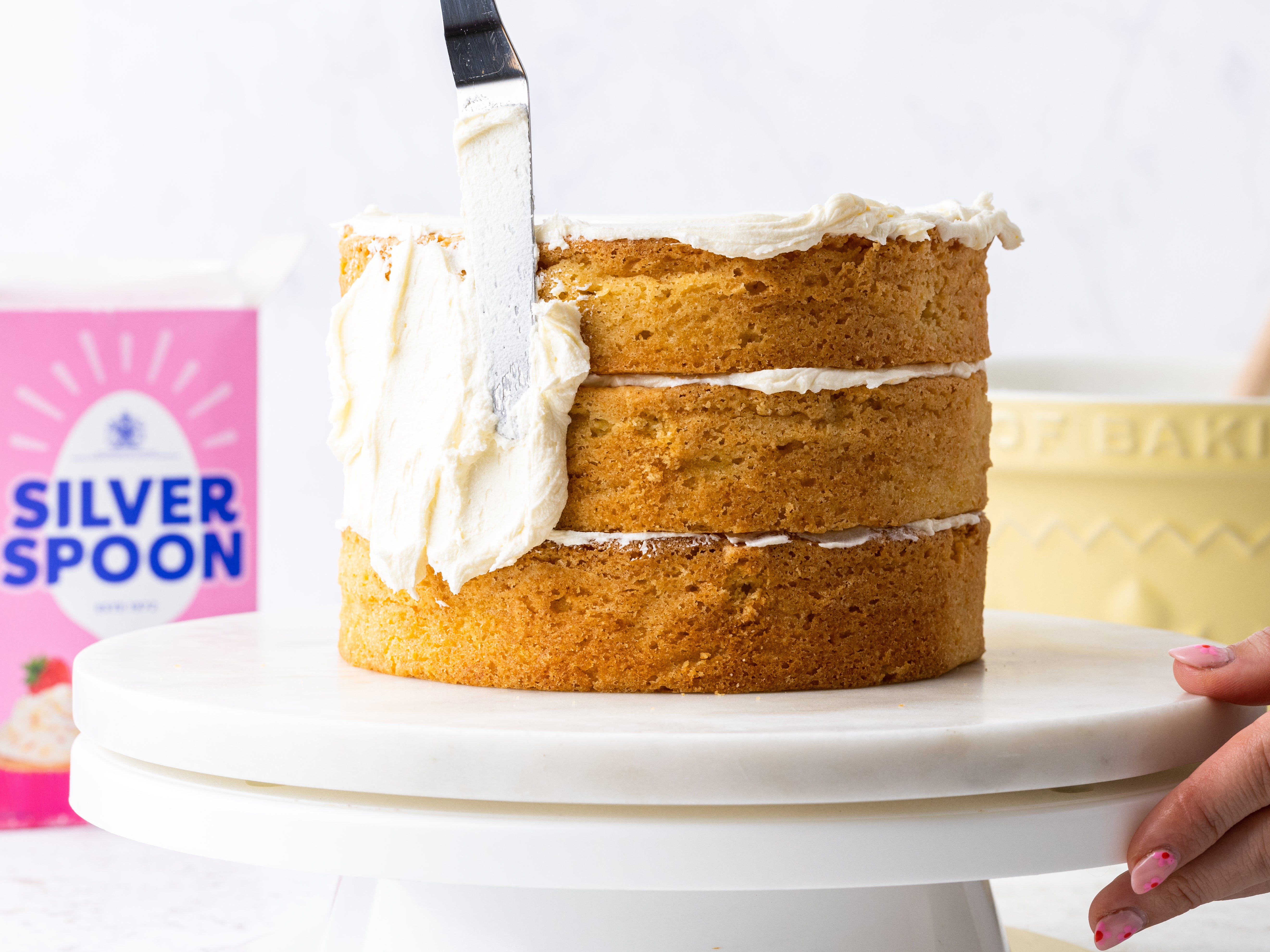 A 3 tied cake being iced on a cake plinth, with a pink packet of Silver Spoon icing sugar in the background.