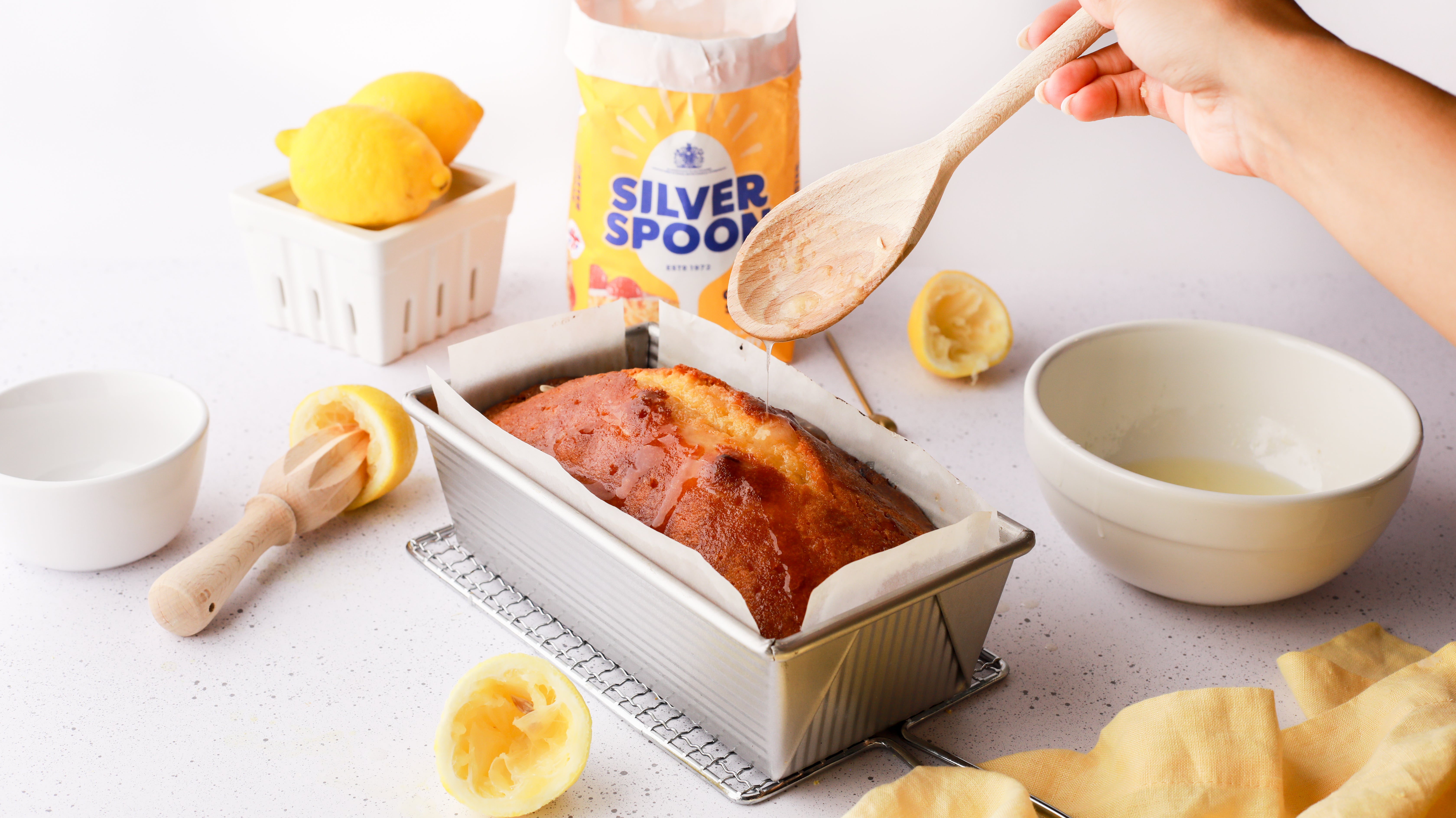 A messy kitchen scene with lemon drizzle glaze being drizzled over a freshly baked lemon cake using a wooden spoon.