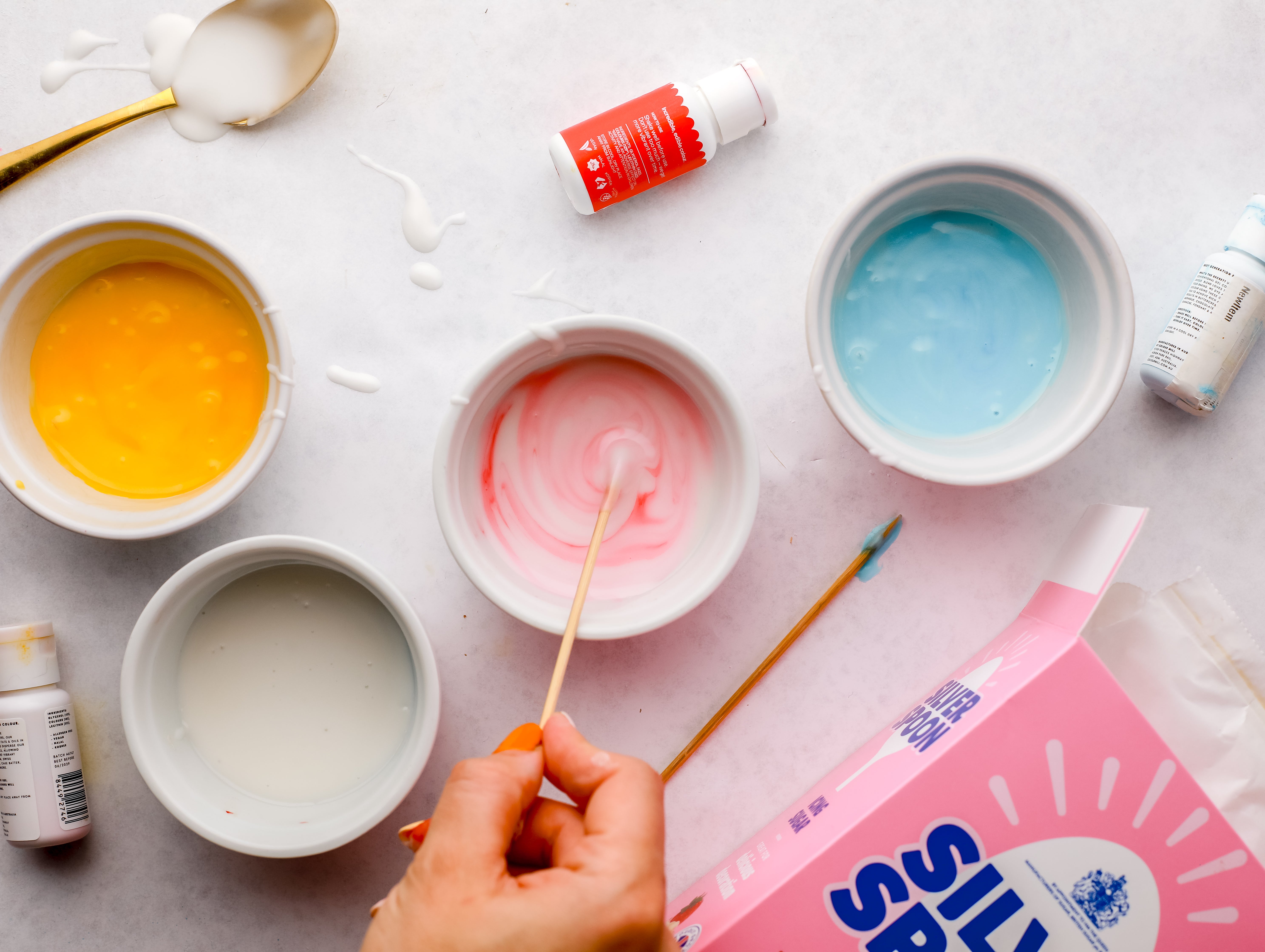 4 round dishes of coloured icing sugar being made up in various blue, pink and yellow colours next to a pink pack of Silver Spoon icing sugar.