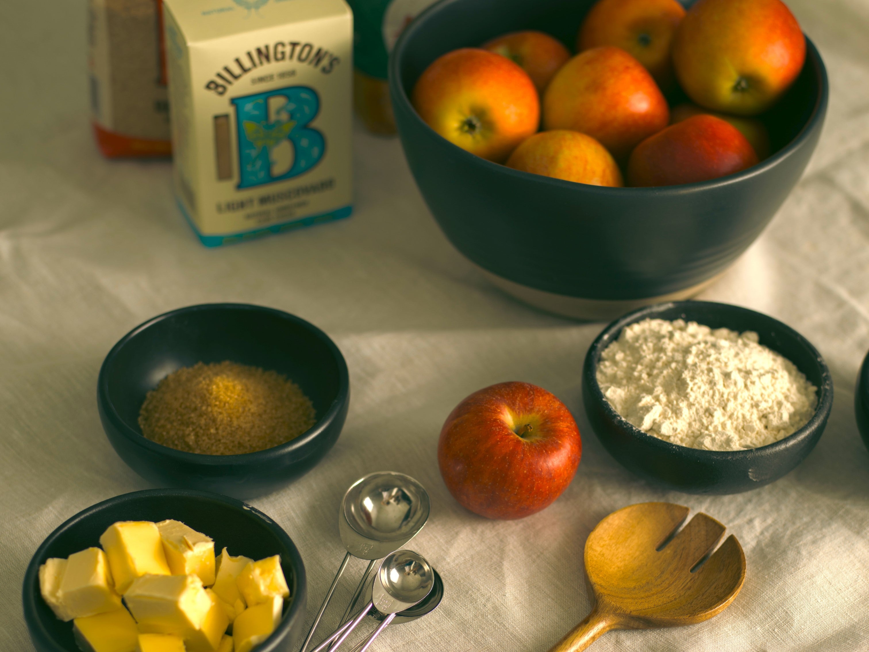 Ingredients for an apple crumble sitting on a white tablecloth next to a box of Billington's sugar