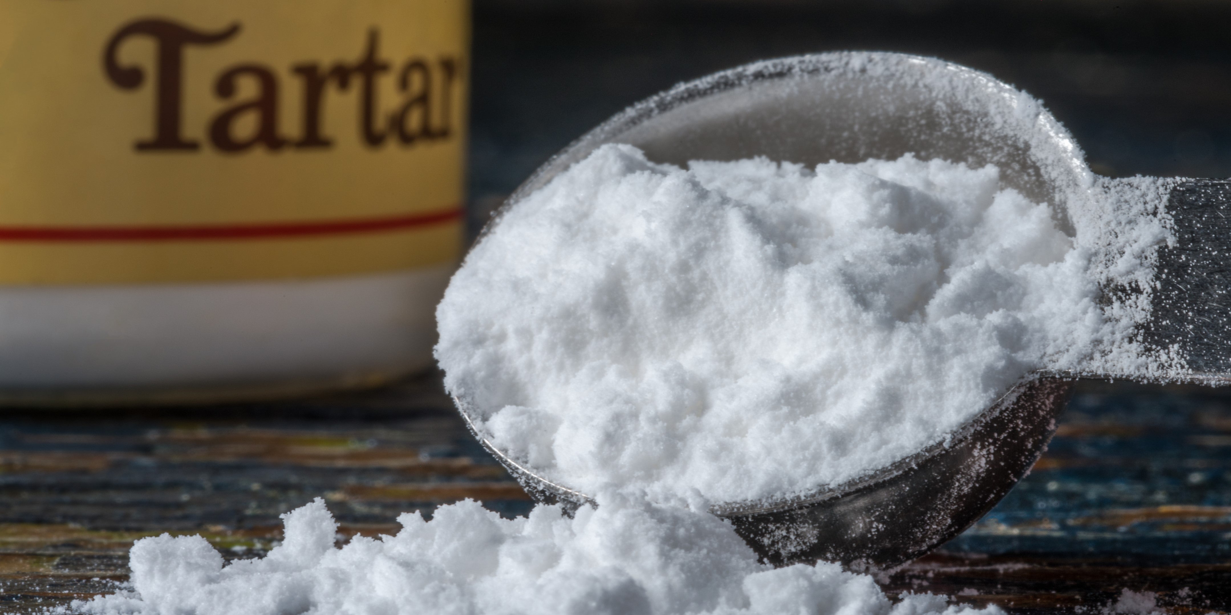 Cream of tartar powder being spilled from a metal spoon with the jar in the background