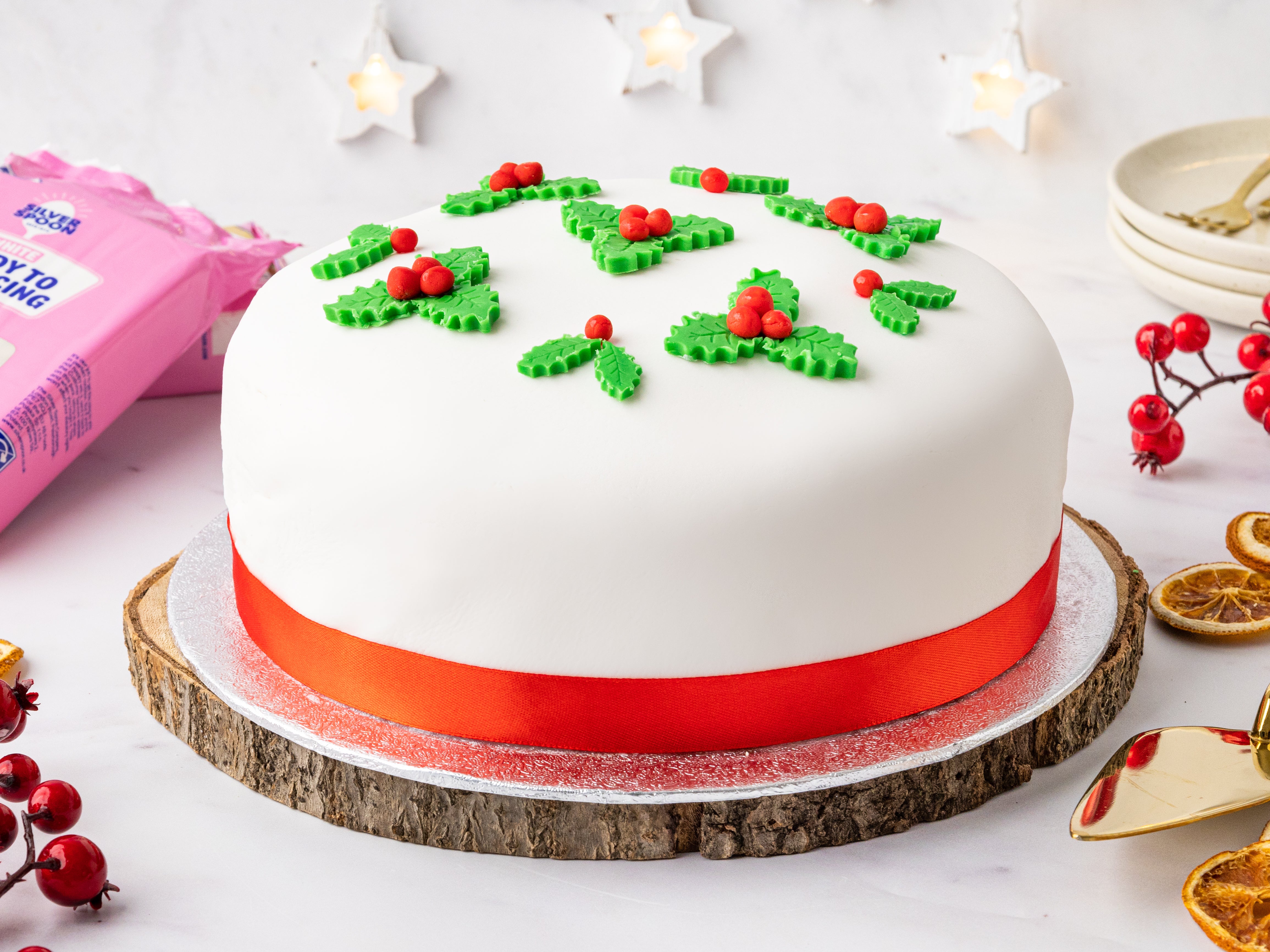 A Christmas cake decorated with white icing and mini icing holly leaves next to a pink pack of Silver Spoon ready to roll icing sugar.
