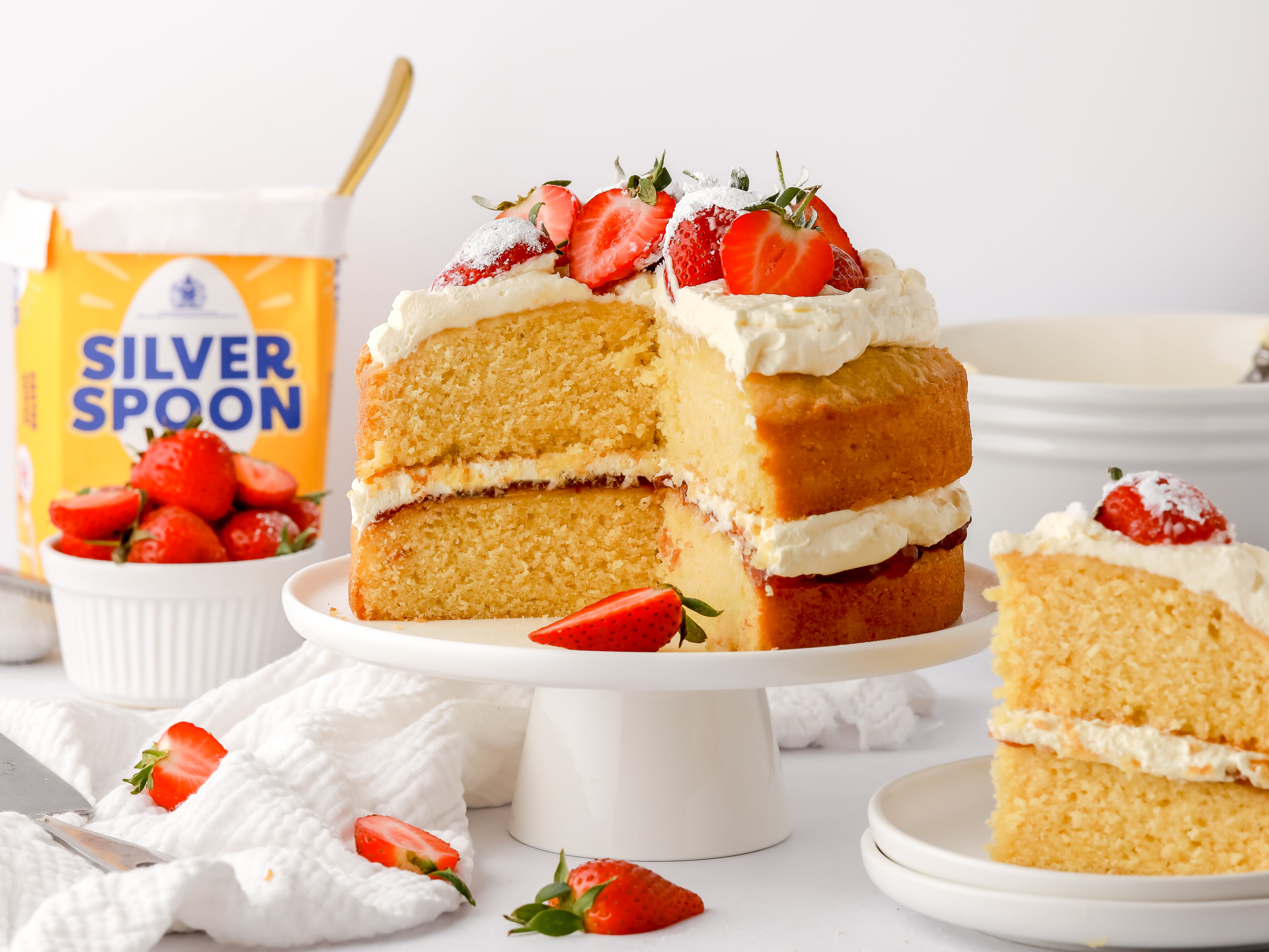 A freshly made Victoria Sponge cake sat on a cake plinth, topped with strawberries and buttercream next to a yellow pack of Silver Spoon caster sugar.