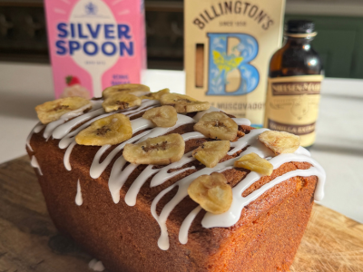 Banana Bread loaf on a wooden chopping board drizzled with icing and topped with banana slices, infront on a vanilla extract bottle and sugar packs