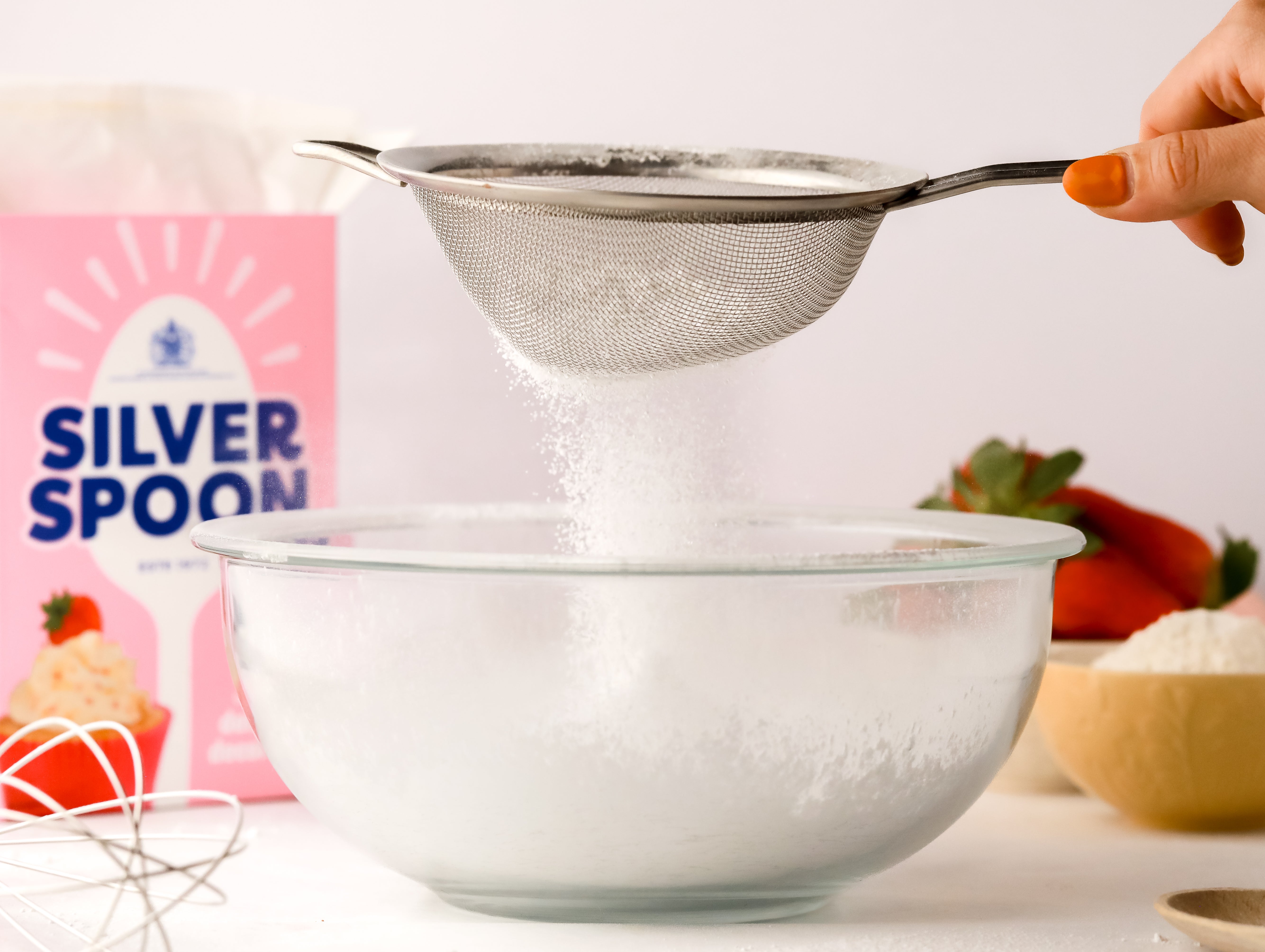 Icing sugar being sieved into a glass baking bowl with a pink pack of Silver Spoon icing sugar in the background, amongst a messy kitchen scene.