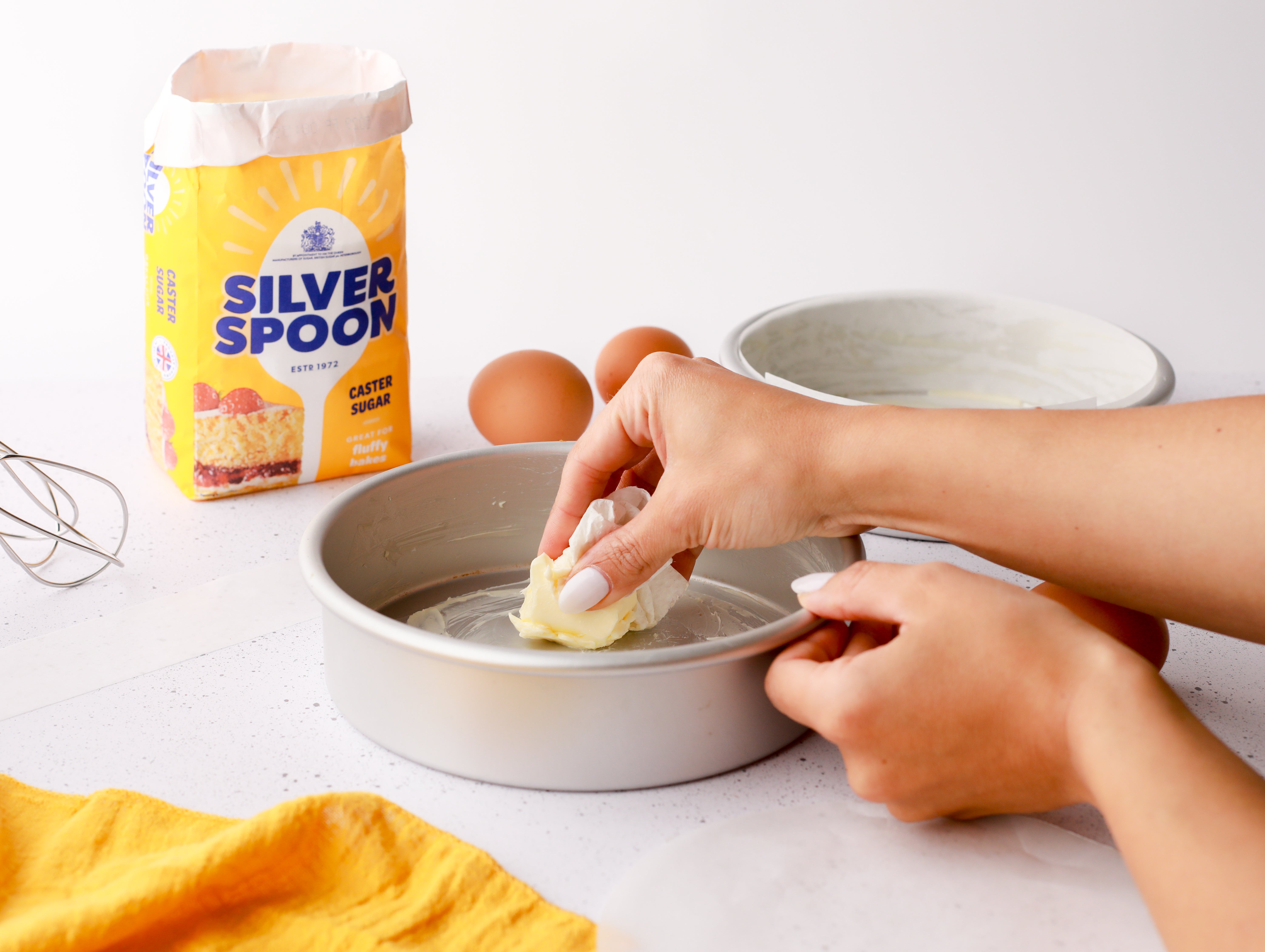 A yellow pack of Silver Spoon caster sugar next to an out of shot person greasing a round baking tin with butter.
