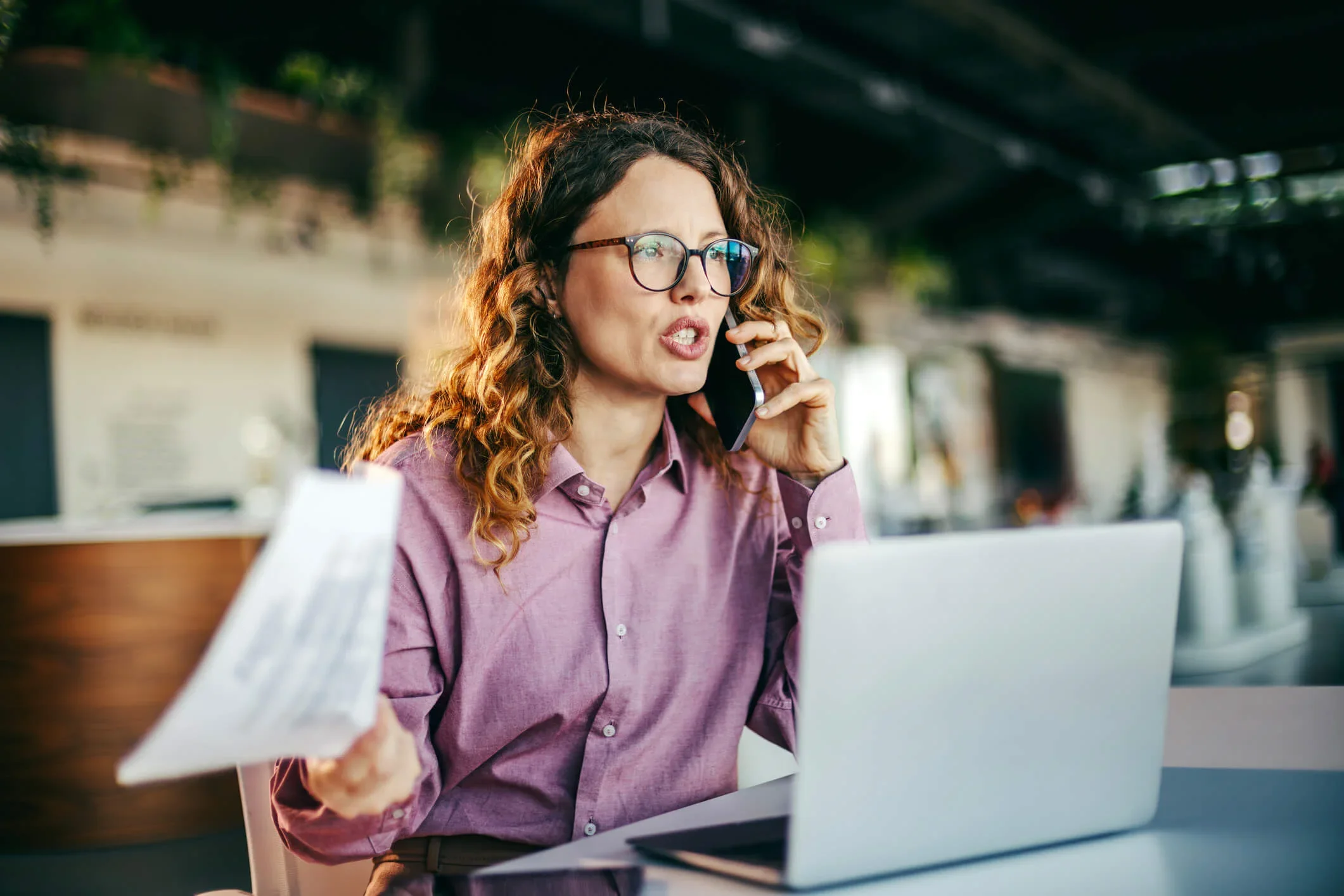 business owner wearing glasses talks on a smartphone while working on a laptop and holding a business energy bill at a desk while complaining to their energy supplier