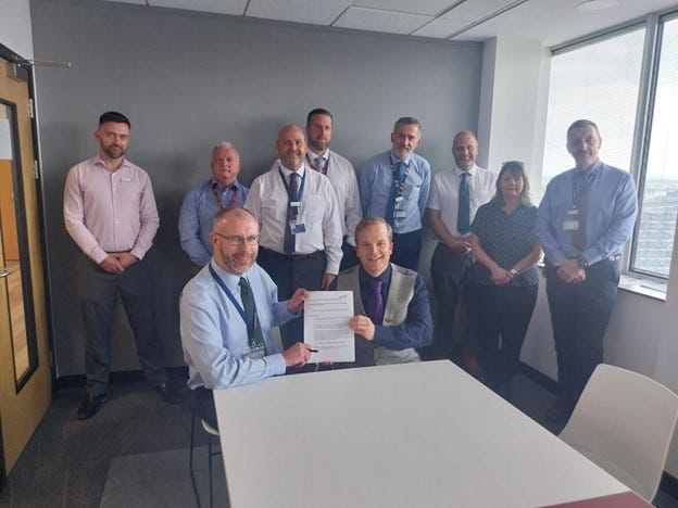 TSSA Organiser Alan Valentine and Transpennine Express Operations Director Rob Cox seated at a table holding up a recognition agreement together. Behind them are a group of men and one woman wearing blue and white shirts. The sun is streaming in through a window to the right.