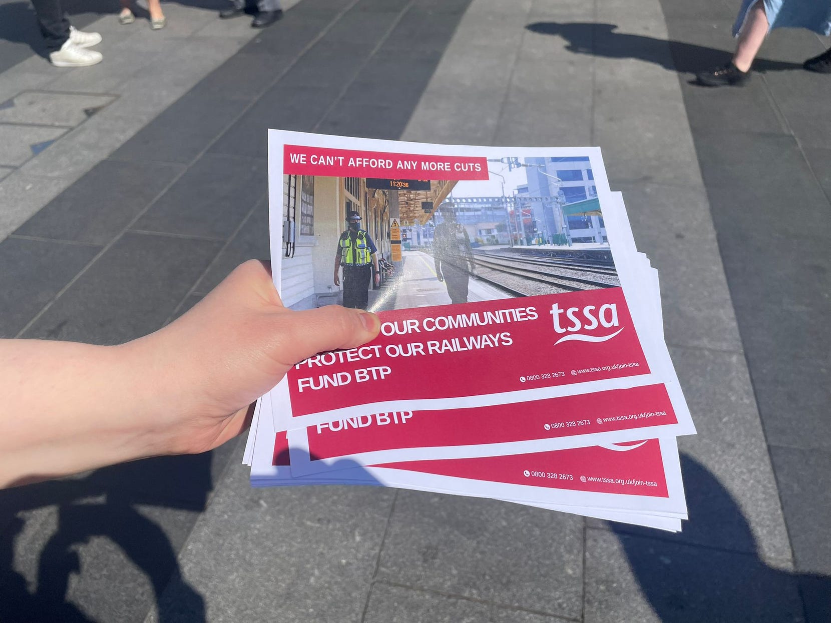 A white hand holds a fan of TSSA leaflets for their campaign to get more funding for the British Transport Police (BTP). The leaflet has a picture of two BTP police officers when one officer has been greyed out so they're just a silhouette.