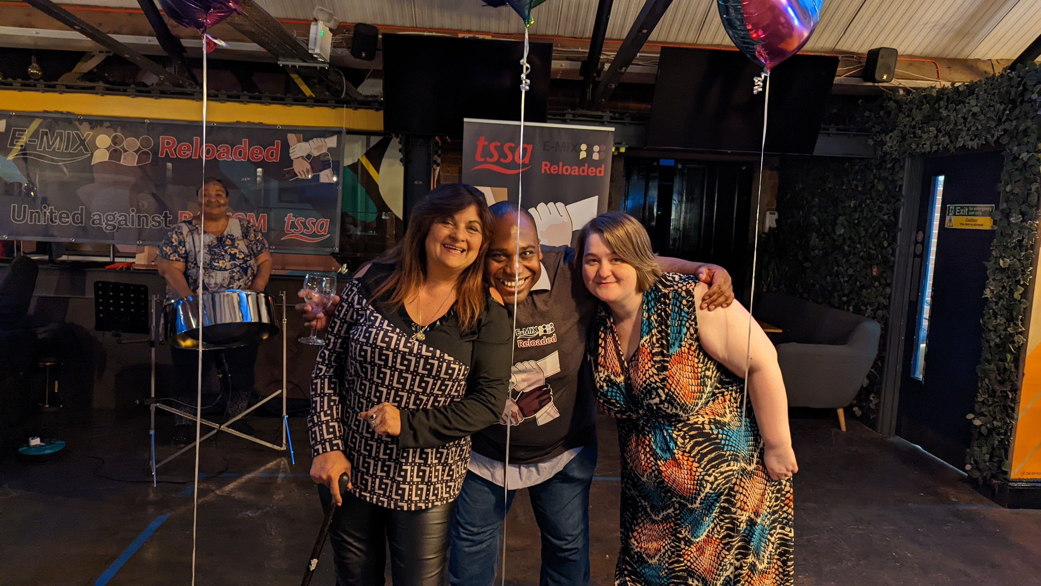An asian woman, a black man and a white woman hugging in a group and holding balloons. There is a black woman playing a steel drum behind them.