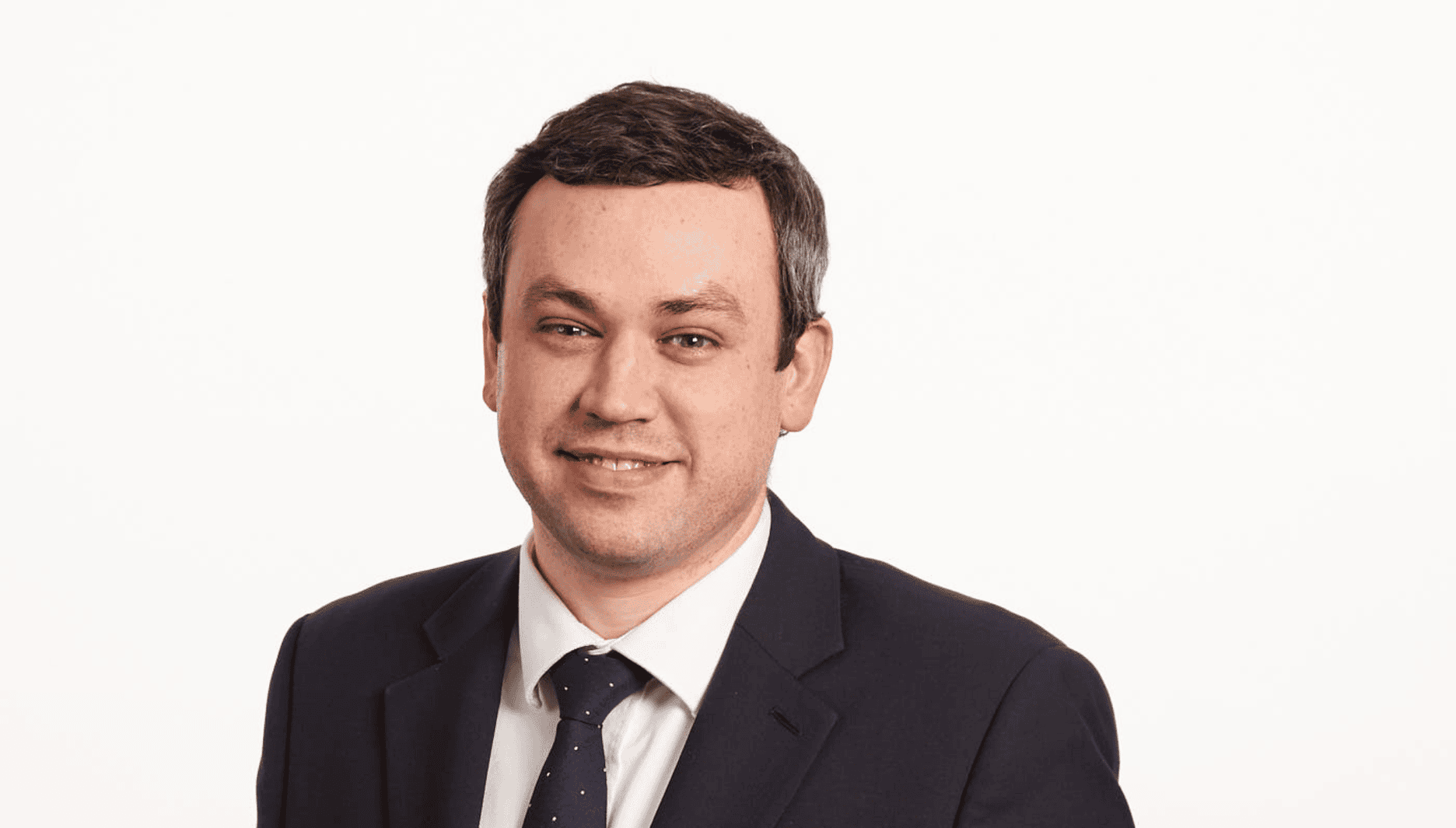 Tony Rippon, a white man, with greying dark hair, in a black suit jacket, white shirt and black tie with tiny polka dots. 