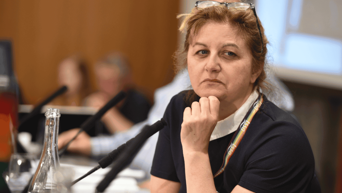 Nicola Jukes, a white woman with brown hair, and her glasses on top of her head. She is sitting in front of a microphone at a conference looking pensive and determined.