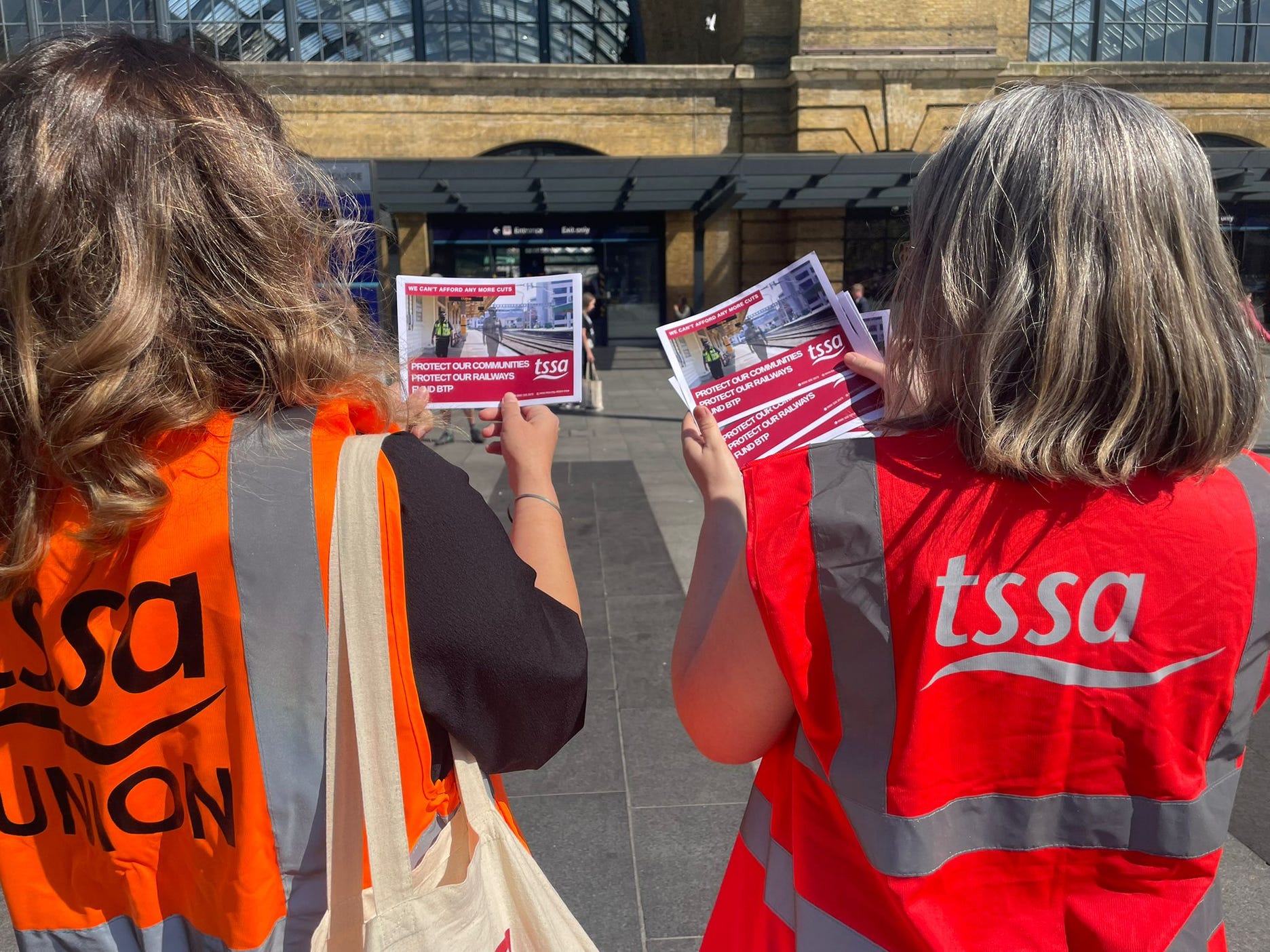 Two women in TSSA hi-vis jackets with their backs to the camera, facing Kings Cross Station. They are holding leaflets for TSSA's campaign to save BTP funding.