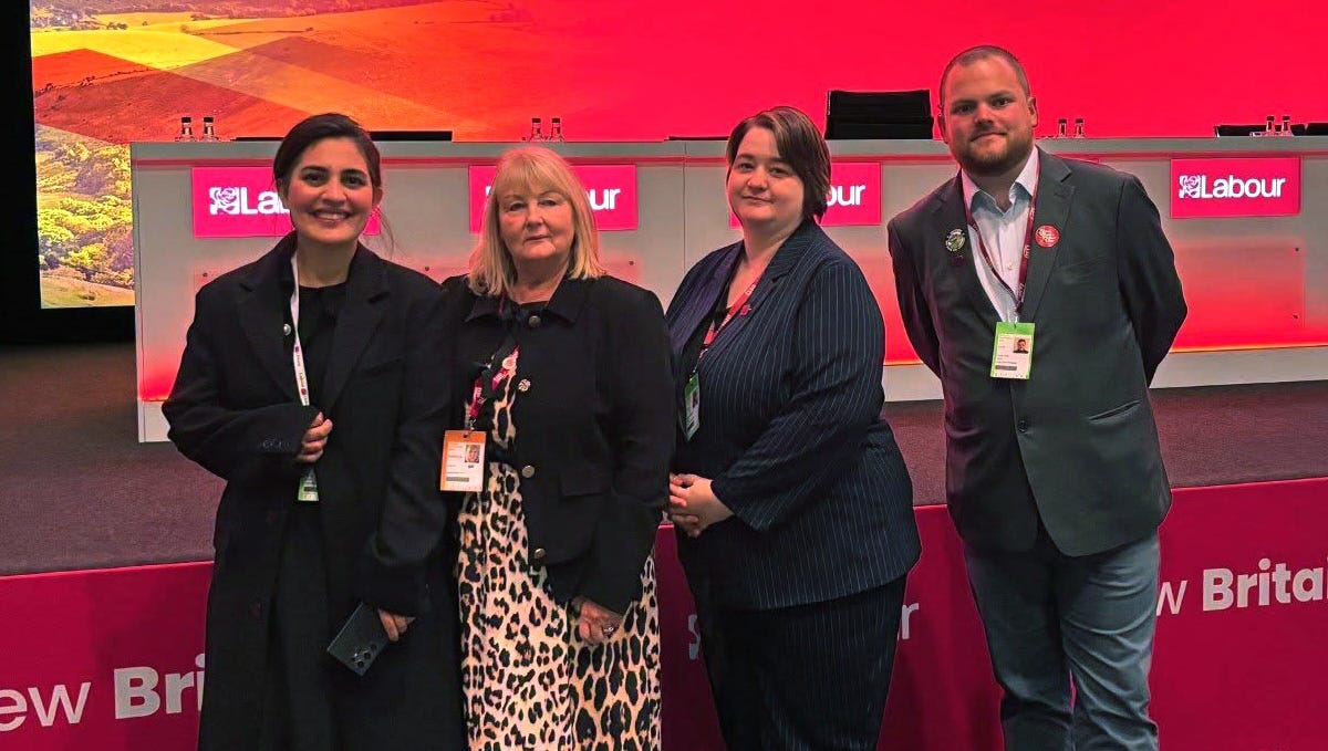Four conference attendees stand together in front of a red-lit stage and registration desk at a Labour Party event. The stage backdrop features a countryside landscape image on a large screen, with Labour branding visible behind the group. All four attendees are wearing event lanyards and badges.
