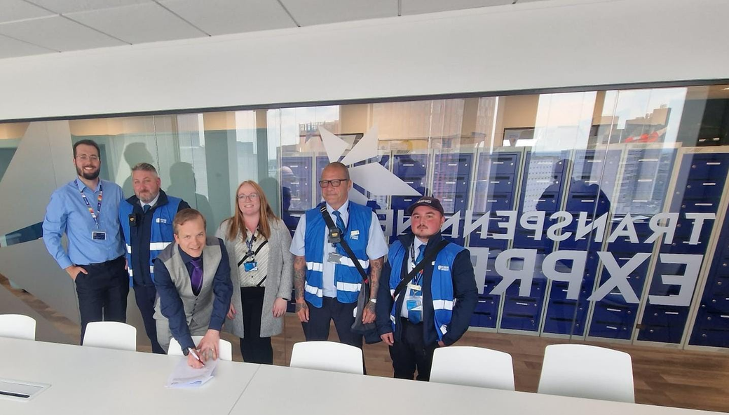 TSSA Organiser Alan Valentine, a short white man, signing a document on a white table. Behind him are five people, men and women, in Transpennine Express uniforms. Behind them is a wall with Transpennine Express written on it, in white on dark blue. 