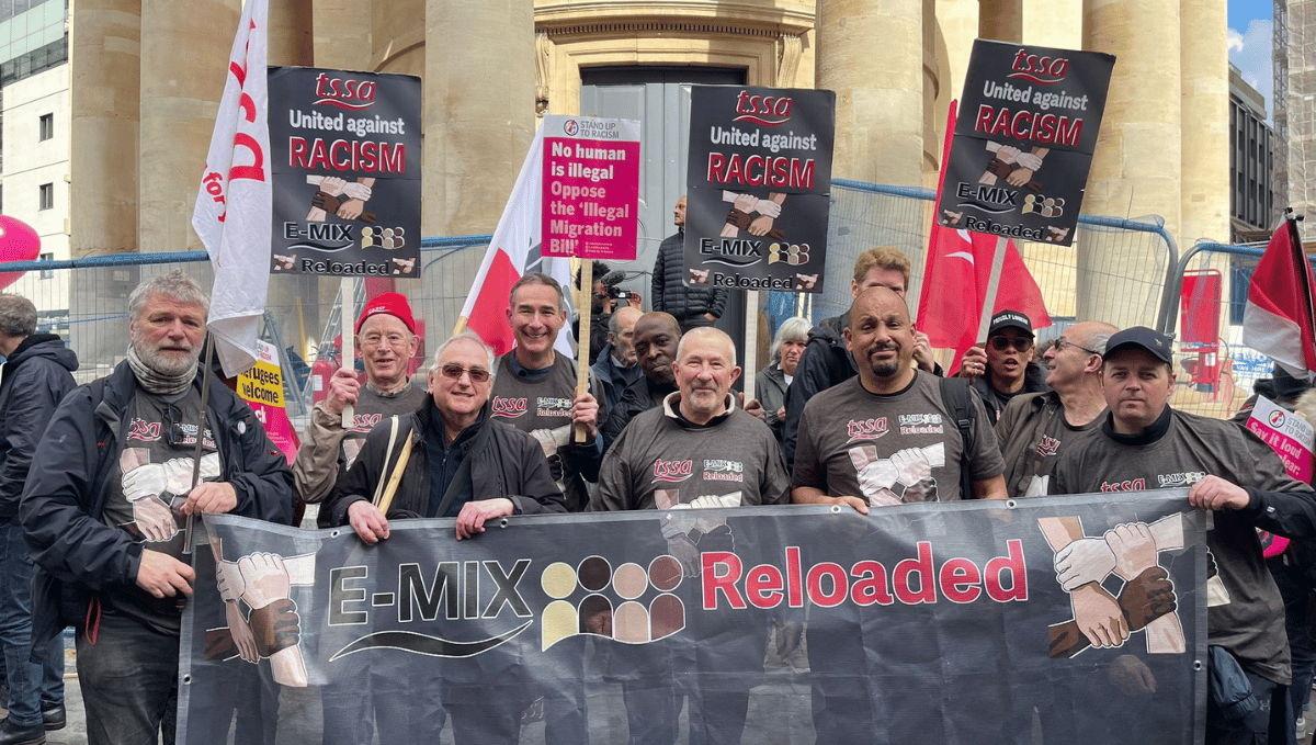 A group of TSSA activists of varying ages and races, wearing TSSA E-mix T-shirts, waving TSSA flags and holding up a big TSSA E-Mix Reloaded banner smile at the camera.