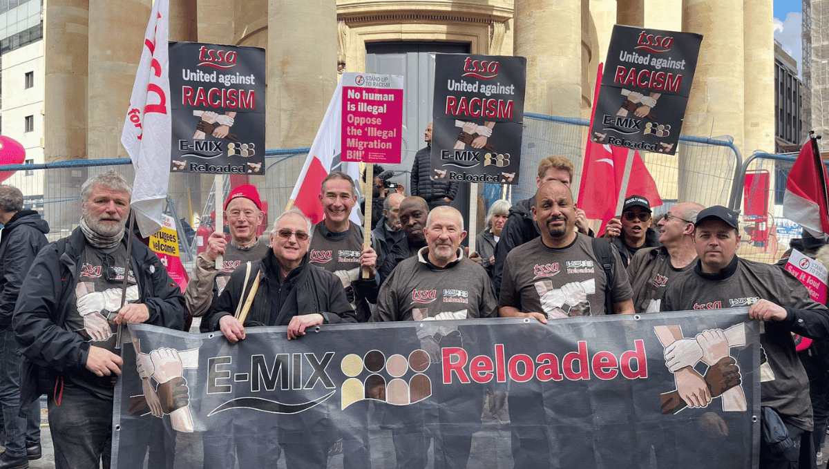 A group of TSSA activists of varying ages and races, wearing TSSA E-mix T-shirts, waving TSSA flags and holding up a big TSSA E-Mix Reloaded banner smile at the camera.