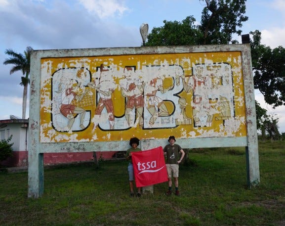Two men standing in front of a big sign which reads 'Cuba', as they hold a red TSSA flag.
