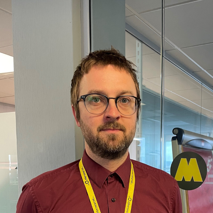 Ian a white man with brown hair and beard, blue eyes, and black framed glasses. He is wearing a maroon shirt with a yellow lanyard. Behind him a yellow Merseyrail "M" logois clearly visible.