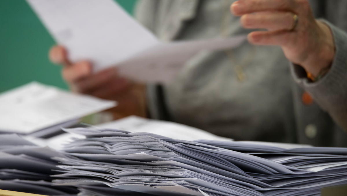A pile of ballot papers with a woman in a grey top sitting behind it holding a another paper.