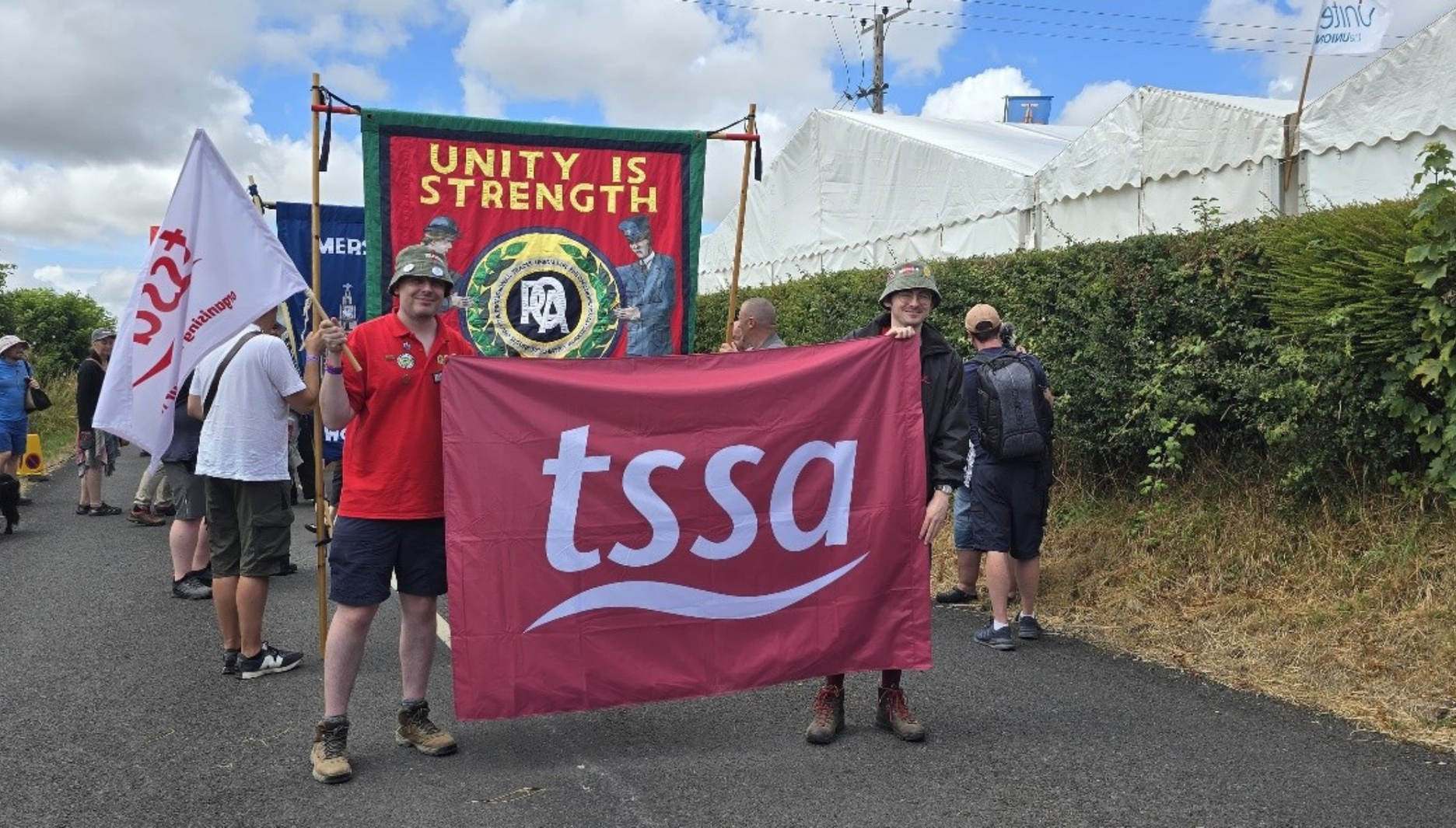 Two TSSA reps are holding a gigantic red flag with a white tssa logo on it. The reps are young white men wearing bucket hats. One is almost hidden by the flag, the other stands to one side. He is wearing shorts and holding a smaller tssa flag with one hand. They are standing in a procession with a big flag saying "Unity is Strength" behind them. 