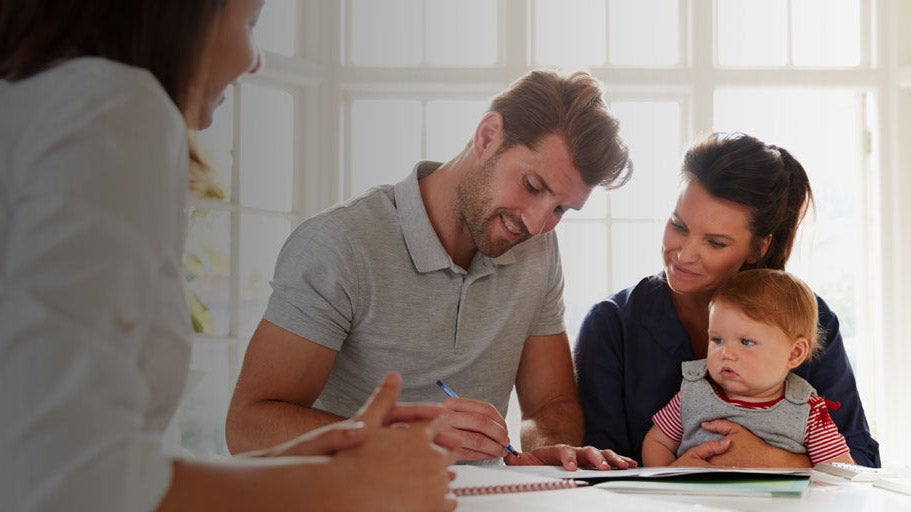 A man and woman sat down with their baby on the mums knee, they are both looking down at paperwork, whilst the man is holding a pen. 